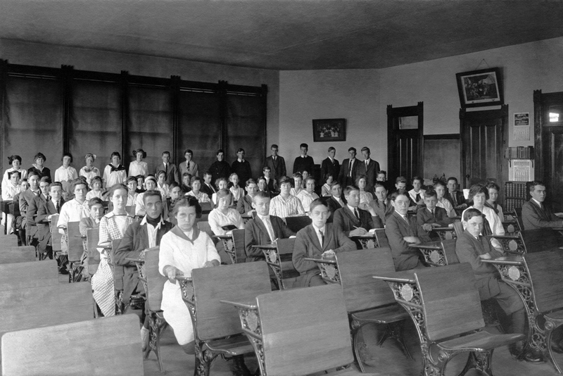 دانش آموزانی که روی میزها و کلاس درس نشسته اند و تعدادی پشت سر هم ایستاده اند. کتیبه بالای عکس کلاس 1919 را می خواند